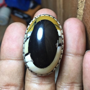 May include: An oval ring featuring a large, polished cabochon stone with a prominent black center surrounded by bands of cream, yellow, and white. The stone is set in a silver-toned metal frame with a textured, notched edge detail around the perimeter.