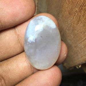 May include: An oval-shaped, translucent gemstone with a smooth, polished surface. The stone displays a milky, pale white color with subtle cloudy inclusions and a soft, reflective sheen across its rounded, cabochon-style top surface.