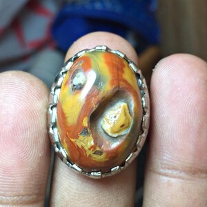 May include: An oval-shaped cabochon gemstone ring featuring a polished surface with swirling patterns of orange, yellow, and brown hues. The stone is set in a silver-toned metal band with a scalloped, notched edge surrounding the perimeter of the setting.