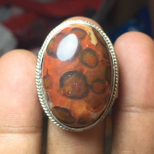 May include: An oval-shaped ring featuring a polished cabochon gemstone with a vibrant orange background and dark brown, circular, leopard-like spots. The stone is set within a silver-toned metal band that has a decorative, twisted rope-style border.