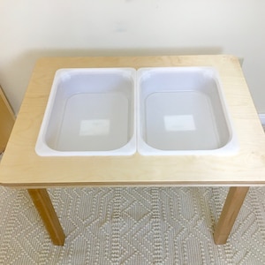 Peut inclure: Une table en bois avec deux bacs en plastique blanc intégrés au dessus. La table a quatre pieds et est posée sur un tapis blanc.