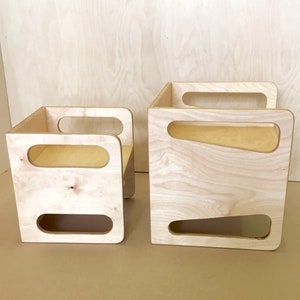 May include: Two unfinished wooden storage cubes. The larger cube has a rectangular shape with a handle on top and a trapezoid-shaped cutout on the side. The smaller cube has a chair-like shape with a handle and a rectangular cutout.