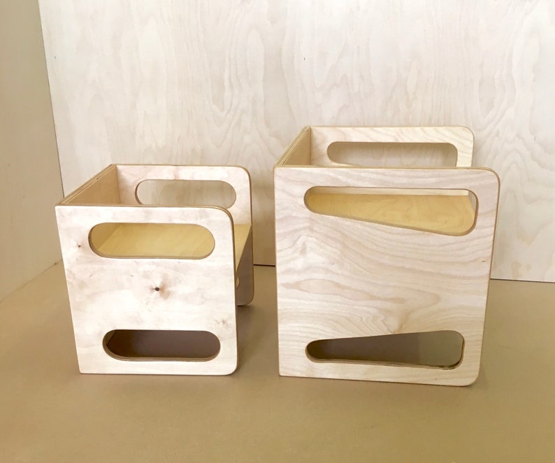 May include: Two unfinished wooden storage cubes. The larger cube has a rectangular shape with a handle on top and a trapezoid-shaped cutout on the side. The smaller cube has a chair-like shape with a handle and a rectangular cutout.