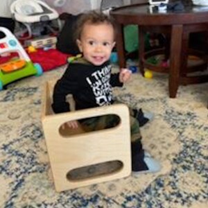 May include: A young child sits on a wooden step stool with two openings. The child is wearing a black long-sleeved shirt with white lettering that says "I think I can I know I will". The child is wearing camouflage pants and has brown hair.