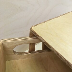 May include: Close-up of a light-colored wooden box with a handle cutout and a lid. The box is made of layered wood, with visible grain patterns. The lid is slightly ajar, revealing the interior of the box.