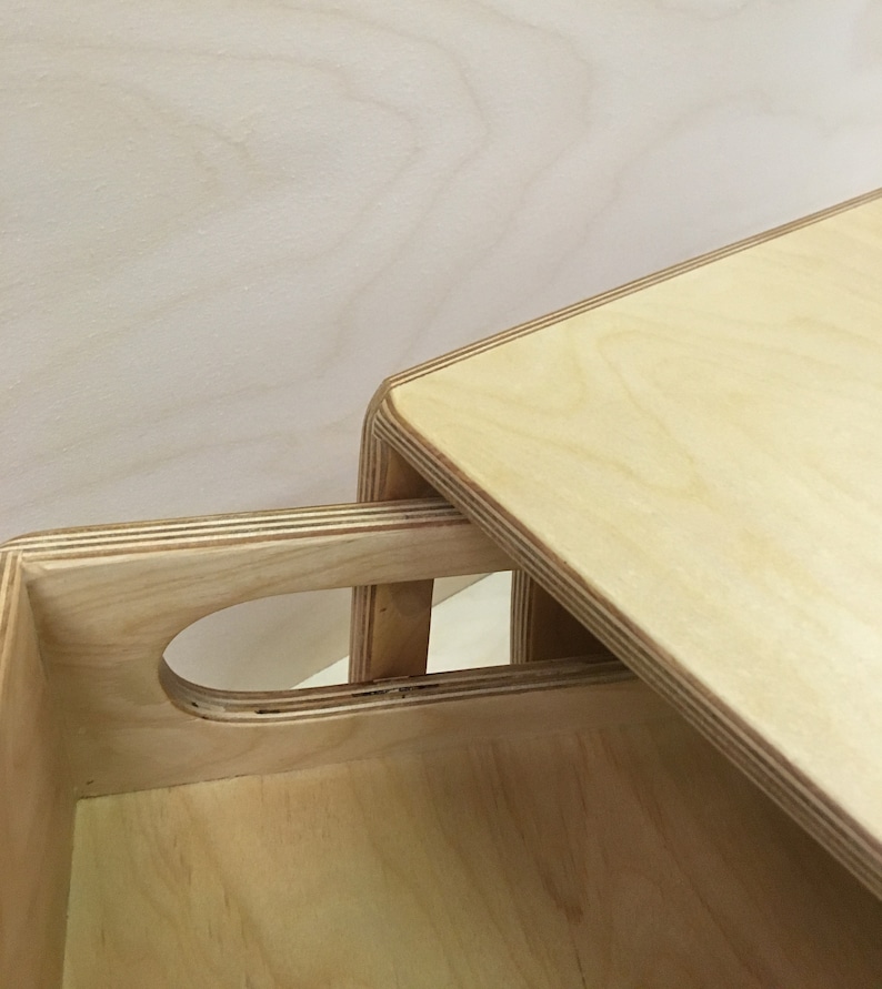 May include: Close-up of a light-colored wooden box with a handle cutout and a lid. The box is made of layered wood, with visible grain patterns. The lid is slightly ajar, revealing the interior of the box.