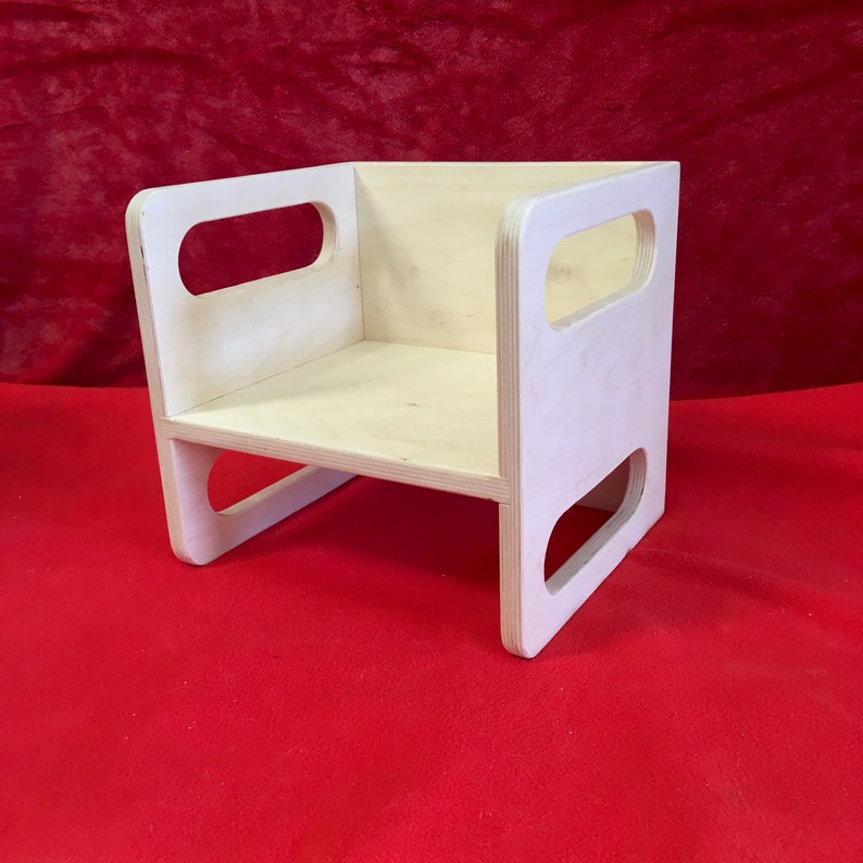 May include: A simple, unfinished wooden chair with a rectangular seat and two cut-out handles on each side. The chair is made of light-colored wood and is sitting on a red surface.
