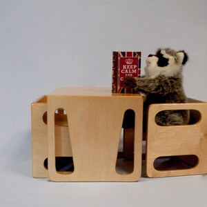 May include: A light-colored wooden table and two matching cube-shaped seats. A stuffed raccoon is sitting on one of the seats, holding a book with the text "KEEP CALM AND CARRY ON". The table and seats are made of light-colored wood.