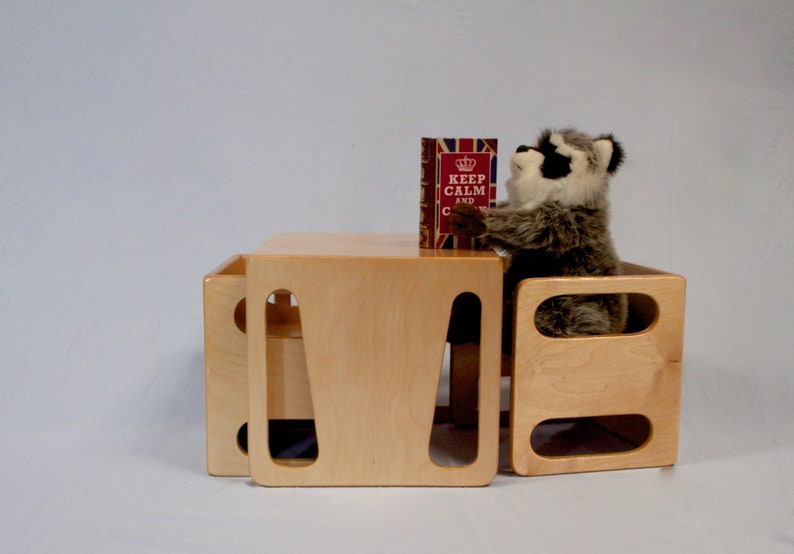May include: A light-colored wooden table and two matching cube-shaped seats. A stuffed raccoon is sitting on one of the seats, holding a book with the text "KEEP CALM AND CARRY ON". The table and seats are made of light-colored wood.