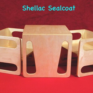 May include: Three wooden, light-colored, open-sided play furniture pieces. The central piece is a cube, flanked by two U-shaped pieces. The text "Shellac Sealcoat" is in the top center. The furniture sits on a red surface.