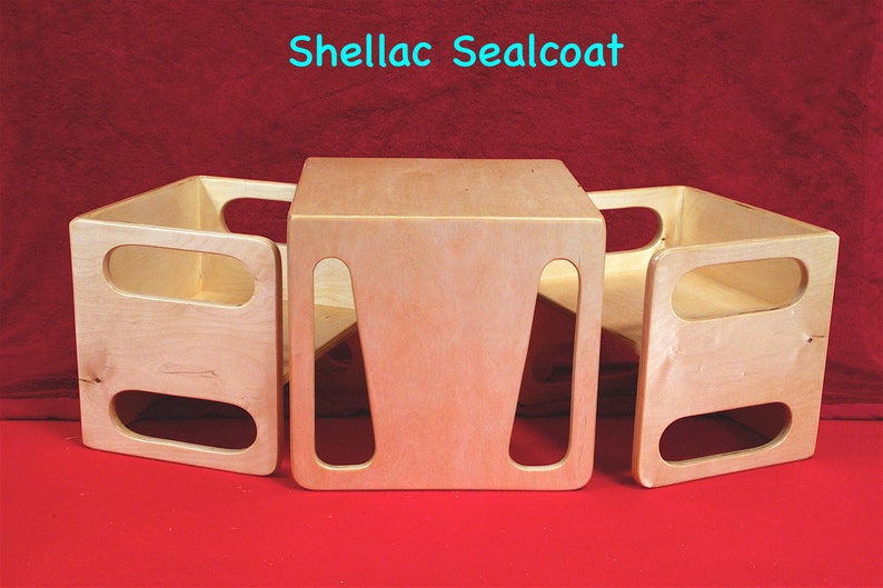 May include: Three wooden, light-colored, open-sided play furniture pieces. The central piece is a cube, flanked by two U-shaped pieces. The text "Shellac Sealcoat" is in the top center. The furniture sits on a red surface.