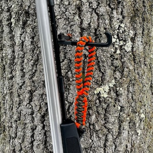 May include: A close-up of a rifle with a black and orange paracord sling attached to the stock. The rifle is leaning against a tree trunk.