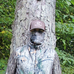 Puede incluir: Una persona con una chaqueta de camuflaje y una m&aacute;scara facial, de pie frente a un &aacute;rbol. La persona lleva una gorra de b&eacute;isbol marr&oacute;n con el texto "EZ Outdoors" en ella.