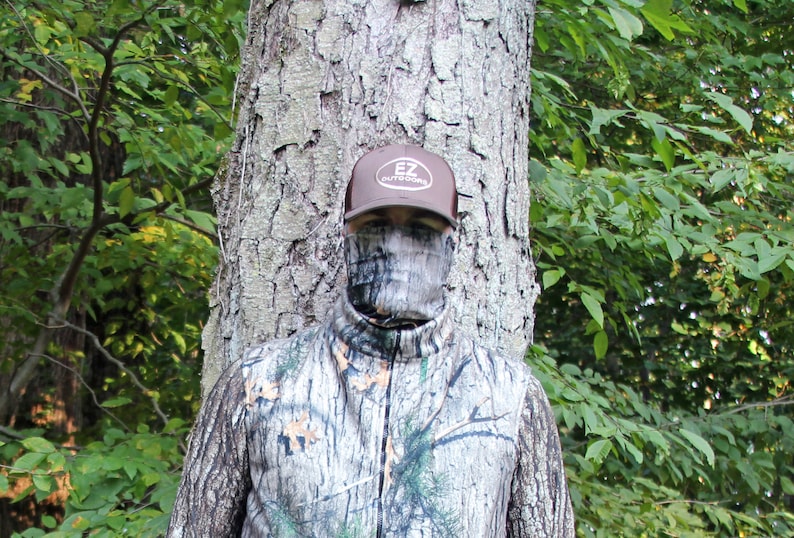 Puede incluir: Una persona con una chaqueta de camuflaje y una m&aacute;scara facial, de pie frente a un &aacute;rbol. La persona lleva una gorra de b&eacute;isbol marr&oacute;n con el texto "EZ Outdoors" en ella.