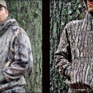 May include: Two men wearing camouflage hoodies. The hoodies are a brown and green camouflage pattern. The men are standing in front of trees.