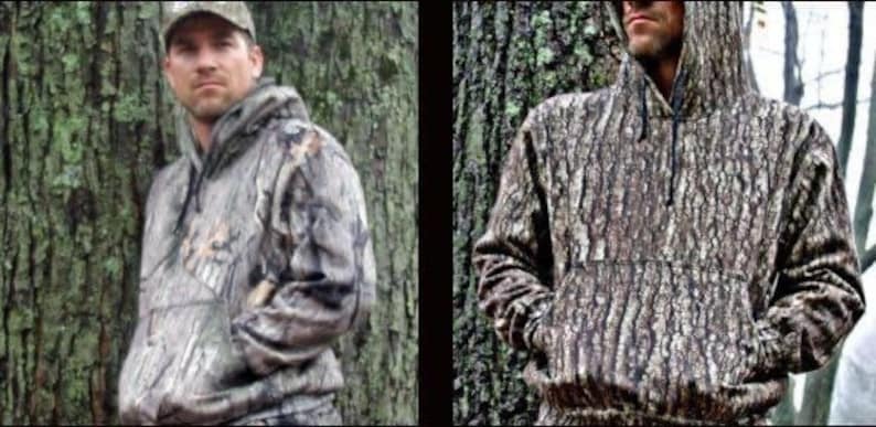 May include: Two men wearing camouflage hoodies. The hoodies are a brown and green camouflage pattern. The men are standing in front of trees.