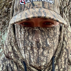Puede incluir: Una persona con una sudadera con capucha de camuflaje y una gorra de b&eacute;isbol de camuflaje a juego que dice "ULTIMATECAMO" en la parte delantera. La persona lleva una m&aacute;scara facial de camuflaje.