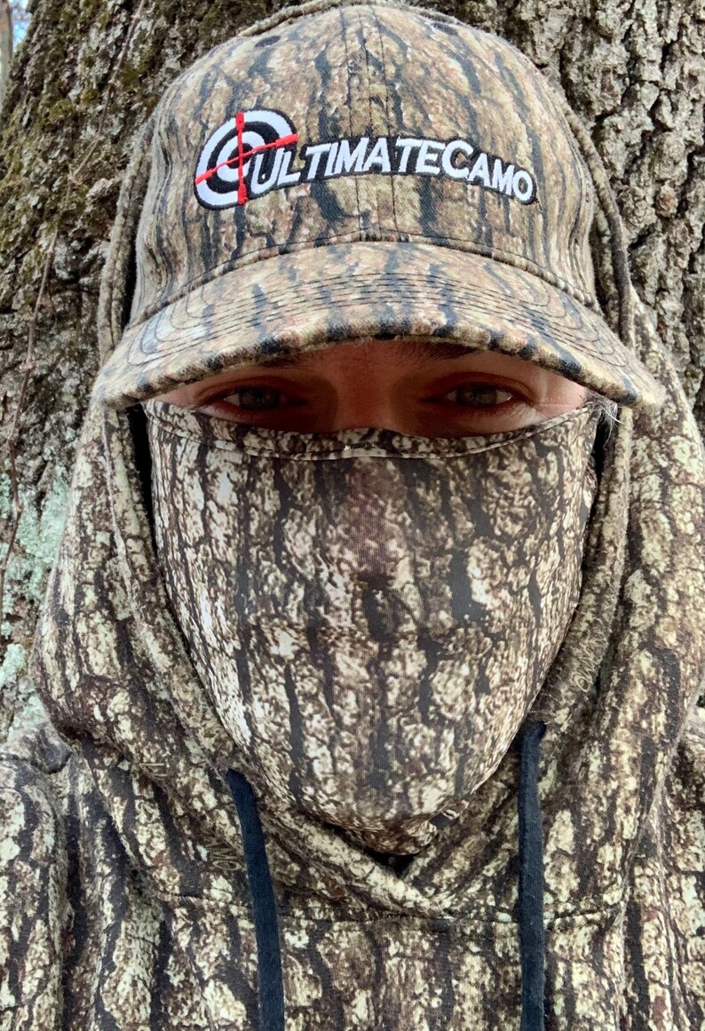 Puede incluir: Una persona con una sudadera con capucha de camuflaje y una gorra de b&eacute;isbol de camuflaje a juego que dice "ULTIMATECAMO" en la parte delantera. La persona lleva una m&aacute;scara facial de camuflaje.