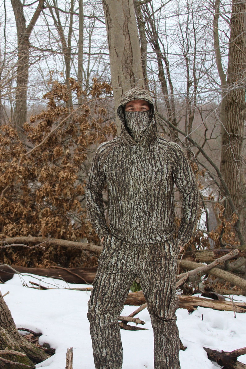 May include: A person wearing a full-body camouflage suit with a tree bark pattern. The suit has a hood that covers the person's head and face. The person is standing in a snowy forest.