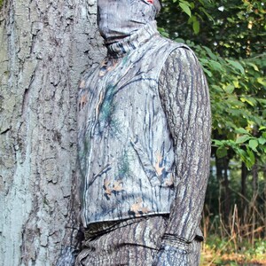 Puede incluir: Una persona con un chaleco de camuflaje y pantalones a juego con una gorra de b&eacute;isbol marr&oacute;n. La persona est&aacute; de pie frente a un &aacute;rbol con un patr&oacute;n de corteza.