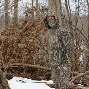 May include: A person wearing a full-body camouflage suit with a tree bark pattern. The suit has a hood with a face mask. The person is standing in a snowy forest setting.