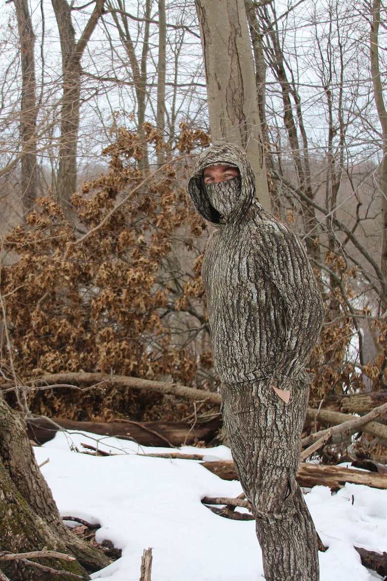 May include: A person wearing a full-body camouflage suit with a tree bark pattern. The suit has a hood with a face mask. The person is standing in a snowy forest setting.