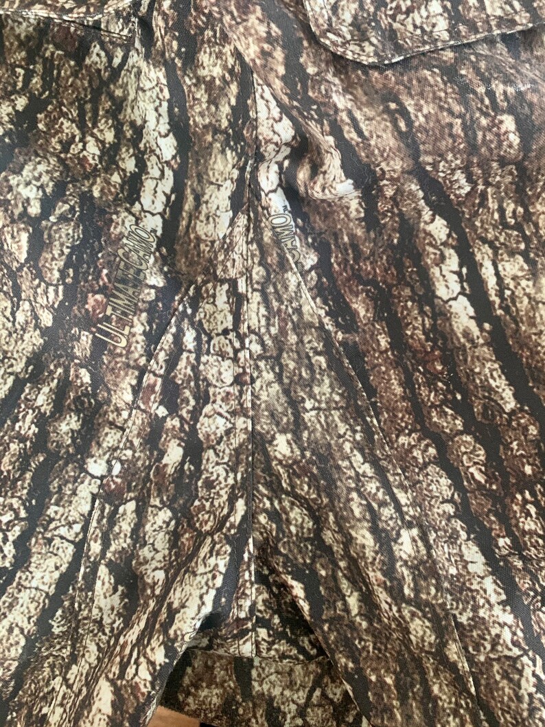 May include: Close-up of a brown and tan camouflage fabric with the text "ULTIMATE CAMO" printed on the left side and "CAMO" printed on the right side.