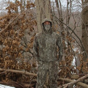 May include: A person wearing a hooded camouflage jacket and pants, standing in a snowy forest setting. The camouflage pattern resembles tree bark.