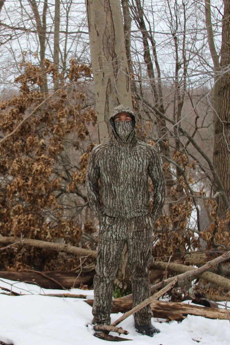 May include: A person wearing a hooded camouflage jacket and pants, standing in a snowy forest setting. The camouflage pattern resembles tree bark.