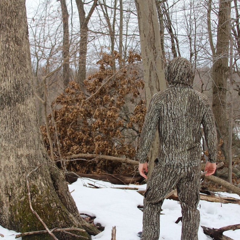 May include: A person wearing camouflage clothing stands in a snowy forest. The person is facing away from the camera and is looking at something in the distance. The forest is covered in snow and there are trees in the background.