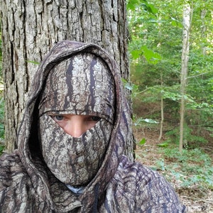 Puede incluir: Una persona con una sudadera con capucha de camuflaje y una m&aacute;scara facial con un patr&oacute;n de corteza. La persona est&aacute; de pie en un bosque.