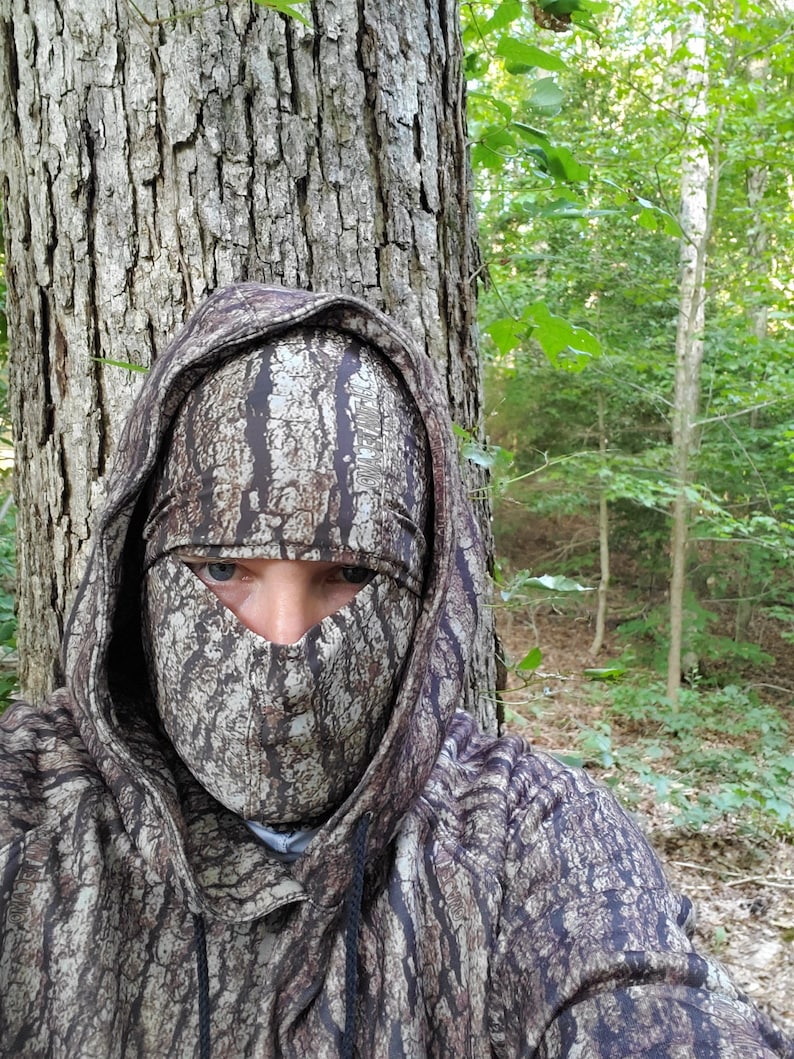 Puede incluir: Una persona con una sudadera con capucha de camuflaje y una m&aacute;scara facial con un patr&oacute;n de corteza. La persona est&aacute; de pie en un bosque.