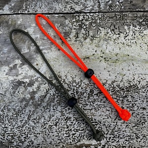 May include: Two adjustable lanyards, one orange and one green, with black plastic adjusters. The lanyards are lying on a weathered wooden surface.