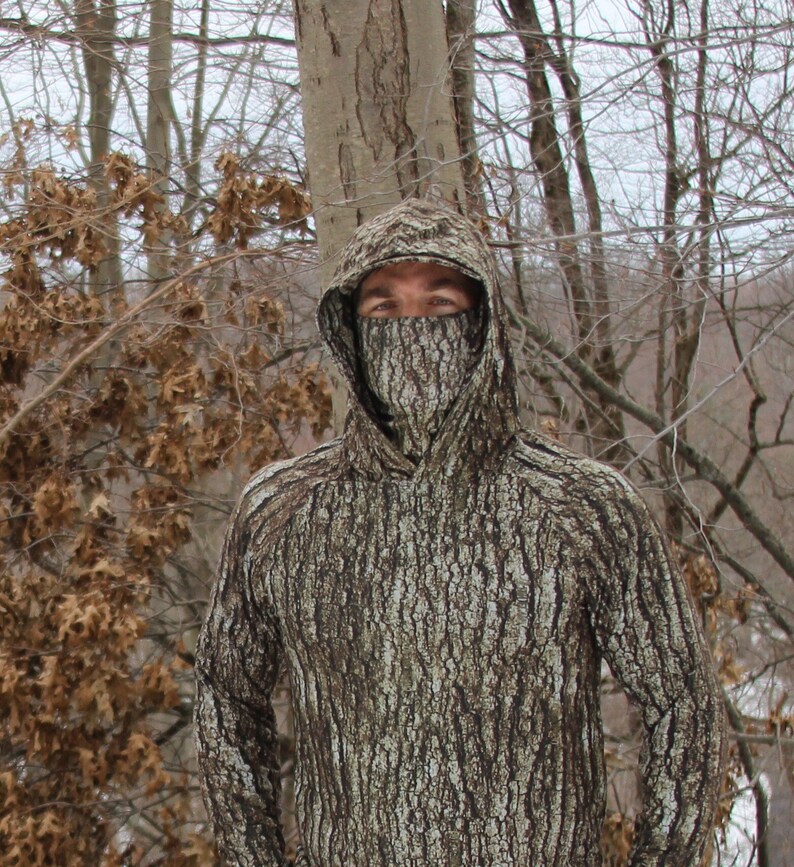 Puede incluir: Una persona con una sudadera con capucha de camuflaje con un patr&oacute;n de corteza de &aacute;rbol. La capucha est&aacute; subida y cubre la cara de la persona.