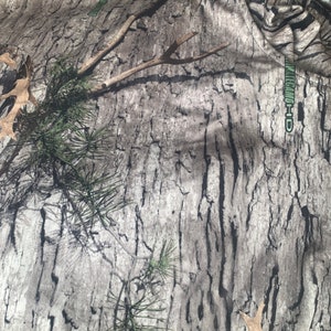 May include: Camouflage fabric with a brown, black, and green pattern. The fabric has the text "REALTREE-HD" printed on it.
