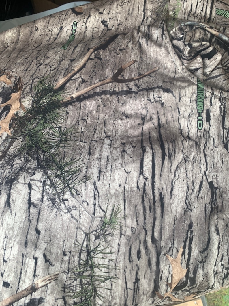 May include: Camouflage fabric with a brown, black, and green pattern. The fabric has the text "REALTREE-HD" printed on it.