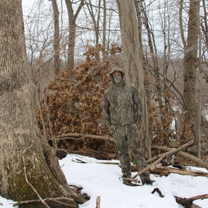 May include: A person wearing camouflage clothing stands in a snowy forest. The person is wearing a hooded jacket and pants with a camouflage pattern. The forest is covered in snow and there are bare trees in the background.