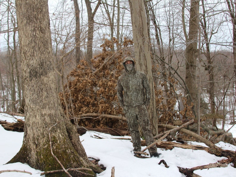 May include: A person wearing camouflage clothing stands in a snowy forest. The person is wearing a hooded jacket and pants with a camouflage pattern. The forest is covered in snow and there are bare trees in the background.