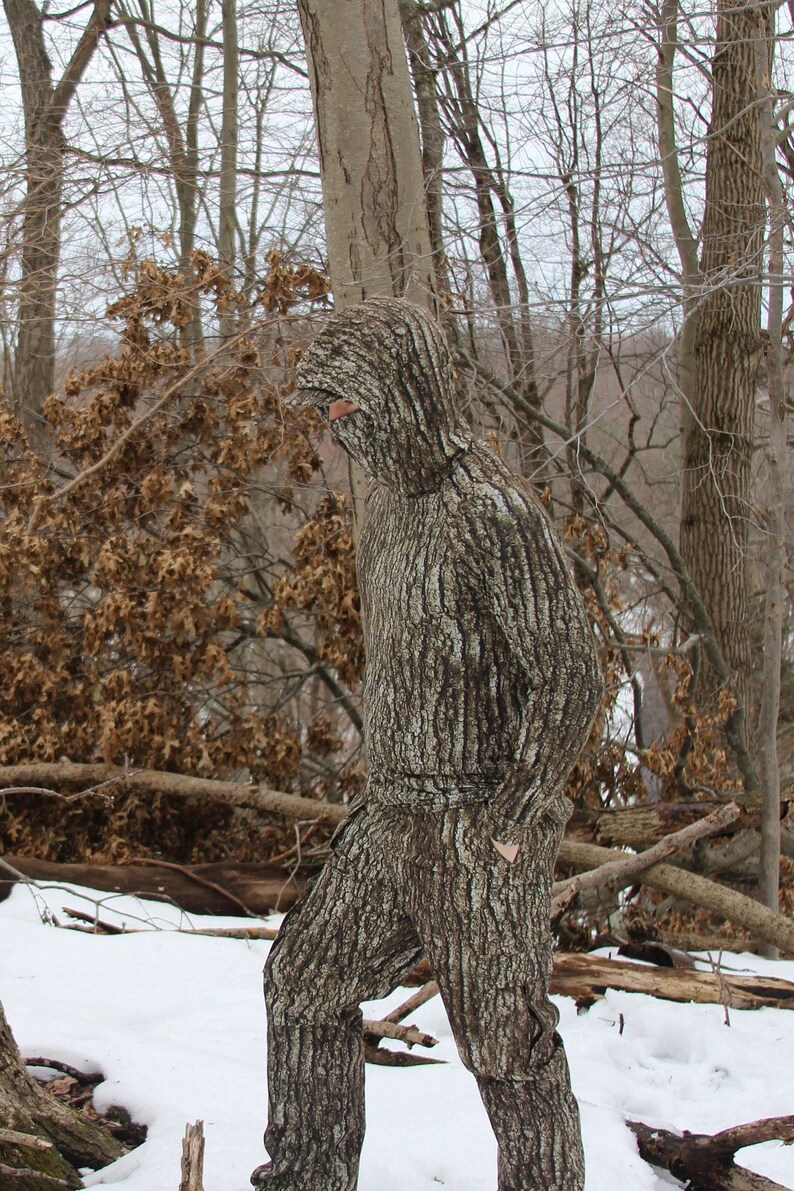 May include: A person wearing a camouflage suit that resembles tree bark, standing in a snowy forest setting.