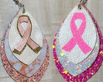 May include: A pair of pink and gold layered dangle earrings with a pink ribbon design. The earrings are made with glitter and faux leather.