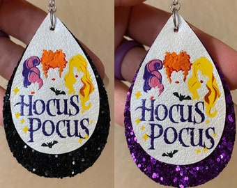 May include: Teardrop-shaped earrings featuring a white top layer with a colorful illustration of three witches and the text "Hocus Pocus". The bottom layer is black glitter on one earring and purple glitter on the other. Silver-tone ear wires.