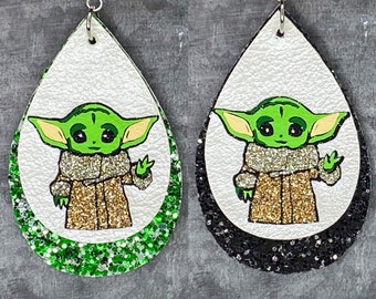 May include: A pair of dangle earrings featuring a green character with large ears and a brown coat on a white background. One earring has a green glitter background and the other has a black glitter background.