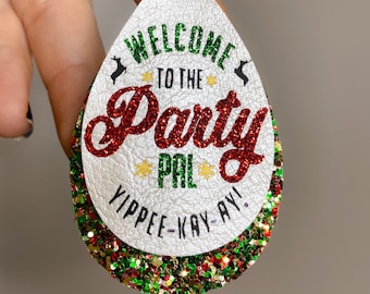 May include: White teardrop-shaped earrings with gold hooks. The earrings are covered in red, green, and gold glitter. The front of the earrings has the text "Welcome to the Party Pal" in red and green lettering. The bottom of the earrings has the text "Yippee-Kay-Ay!" in black lettering.