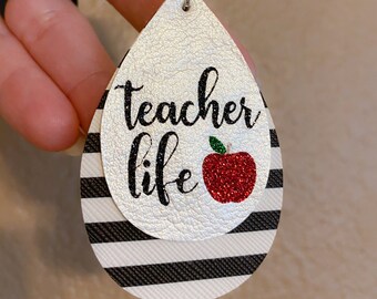 May include: White and black striped teardrop-shaped earrings with a silver glitter background. The earrings have the text "teacher life" and a red glitter apple.