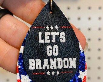 May include: A pair of dangle earrings with a black leather teardrop shape. The teardrop is layered with a red, white, and blue glitter design that resembles the American flag. The text "Let's Go Brandon" is printed in white on the black leather.