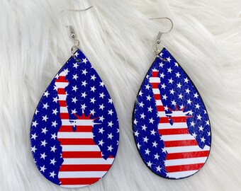 May include: A pair of dangle earrings with a teardrop shape. The earrings are made of leather and feature a red, white, and blue American flag design with a silhouette of the Statue of Liberty.