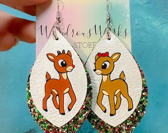 May include: A pair of dangle earrings featuring a white leather base with red and green glitter trim. Each earring has a cartoon image of a reindeer, one with a red bow and the other with a red nose.