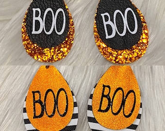 May include: A pair of dangle earrings with a black and orange layered design. The top layer is black leather with gold glitter and the word "BOO" in white. The bottom layer is orange glitter with black stripes and the word "BOO" in black.