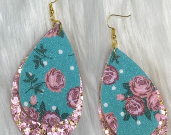 May include: Teal and pink floral dangle earrings with gold hooks. The earrings are made of layered fabric with a glitter finish.