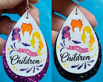 May include: A pair of dangle earrings with a white background and black glitter trim. The earrings feature a graphic of three witches with the text "I Smell Children".